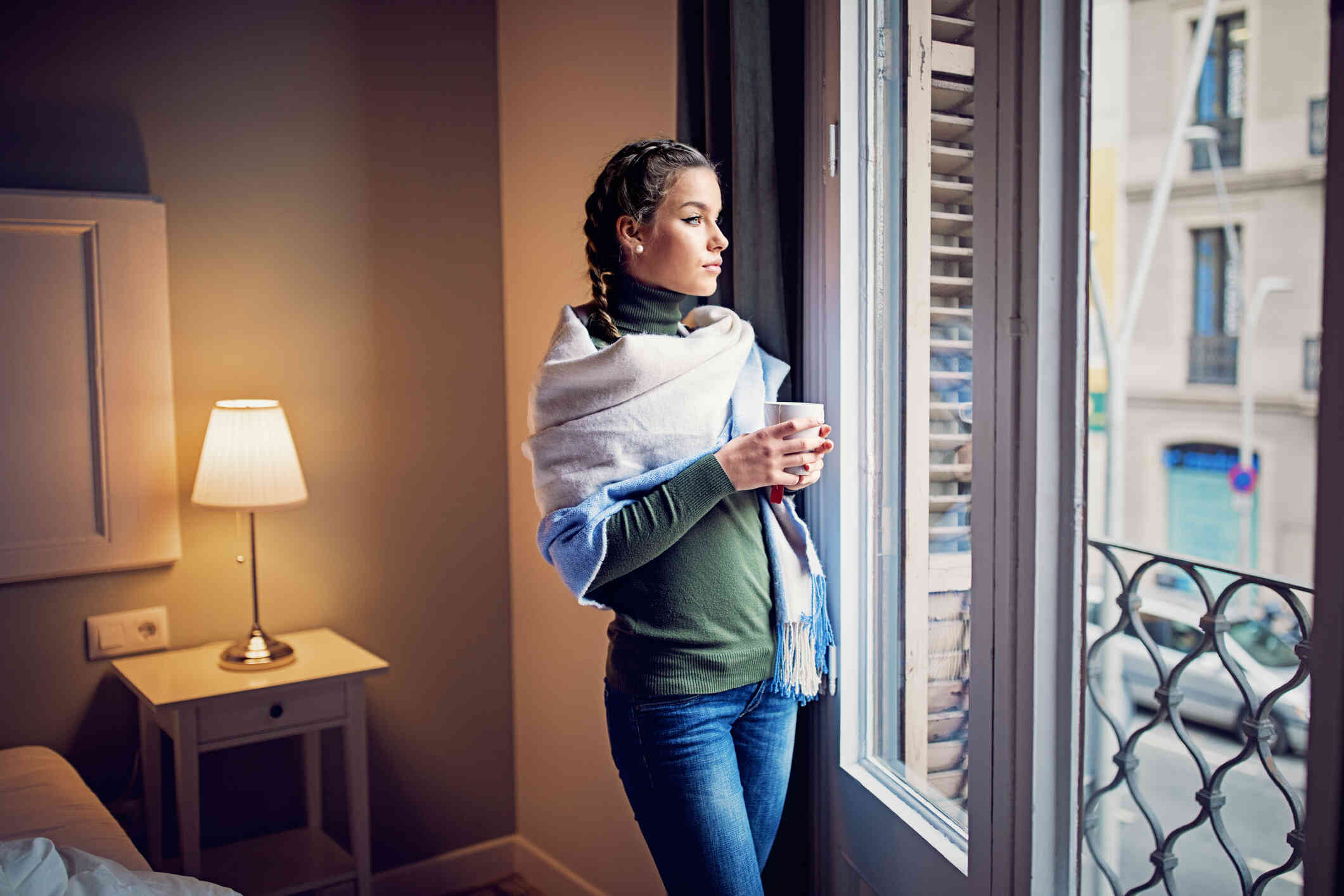 A woman in a shaw holds a mug of coffee while gazing out of the window in her home.